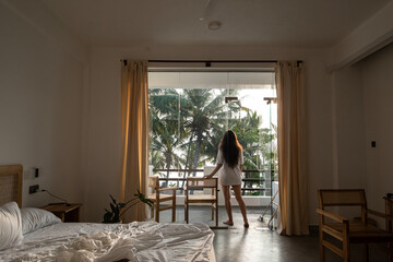 Obraz premium A young woman with long dark hair and a white T-shirt walks out onto the balcony from her luxurious bed after waking up in her hotel room. Wake-up time in a hotel room in Sri Lanka on vacation.