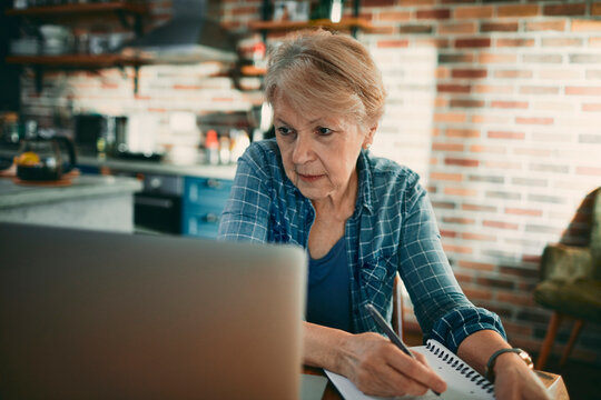 Senior woman taking notes while working online in home kitchen