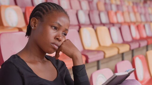 black athlete reviewing tactics on tablet, stadium backdrop, braided hair, focused expression, halftime break, tactical notes, casual training top, empty pastel seats, quiet determination