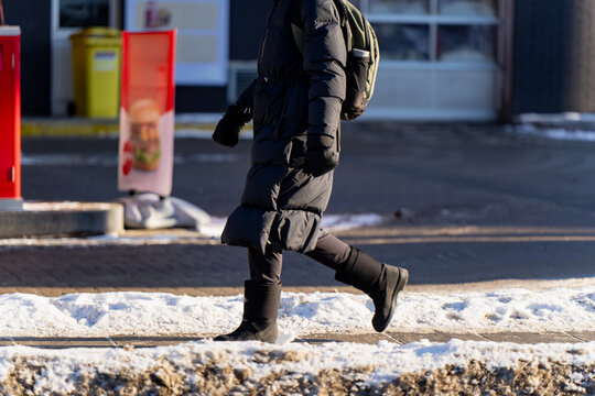 Winter pedestrian crossing snowy sidewalk in black puffer coat and backpack near storefront, urban cold season commute and street safety concept