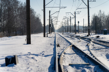 Snowy railway tracks with overhead power lines and poles stretching into distance, winter transport infrastructure corridor under clear blue sky