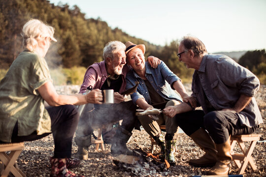 Senior friends cooking fish over campfire on a rocky riverbank