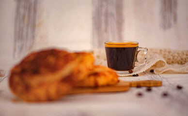 Fresh espresso served in a clear glass cup beside a golden croissant on a wooden board. Soft background blur and warm tones create a cozy caf&eacute; atmosphere with copy space. Selective focus.