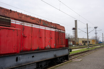 Obraz premium a weathered red train locomotive parked on tracks at an industrial railway station under a grey sky.