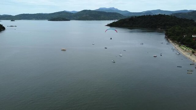 Tandem paramotor flying along Jabaquara Beach and over the water, with a stunning view of the Paraty bay, the small islands and the mountains covered with dense forest in Paraty, Brazil