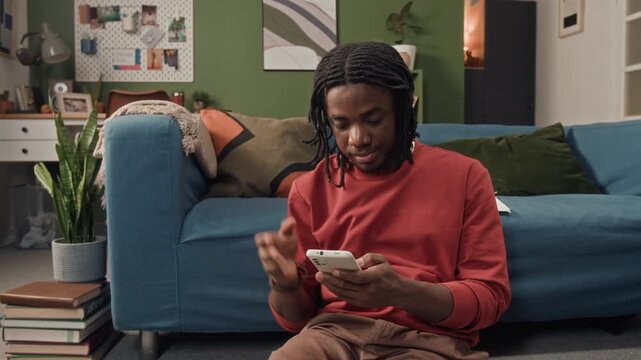 Tired young adult Black man rubbing eyes text messaging on phone while teleworking sitting on floor in living room
