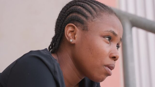 black woman athlete resting on railing, sweat glistening, cornrows braided close, stud earring catching light, contemplative gaze toward distance, breathing steady, mood introspective and determined.