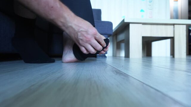 Low angle shot of a man putting on black socks while seated, bare feet on light wood floor, sofa and table blurred behind, capturing quiet daily ritual, comfort, and mindful preparation.