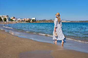 Woman in a light summer dress and straw hat walking barefoot near the shoreline on a Greek beach. Travel and holiday lifestyle scene with coastal town and clear blue sky. © beautiful-photo.de