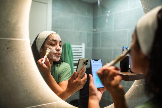 Young woman applying facial mask at bathroom mirror