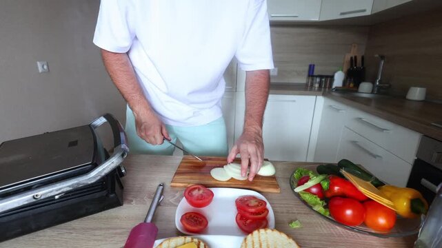 Man slices onion on a wooden board beside a grill press, tomatoes, lettuce, and toasted buns, prepping fresh burger ingredients in a bright kitchen.