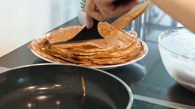 Hands use spatulas to lift thin crepe from hot frying pan and place it onto stack of pancakes. Steam rises from hot pan, cooking process