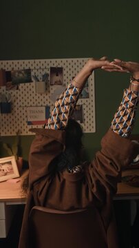 Vertical rear view of young woman taking stretch break extending arms overhead while using laptop at home workstation in dimly lit room