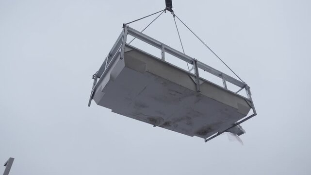 Large industrial metal platform suspended by crane cables against overcast gray sky 