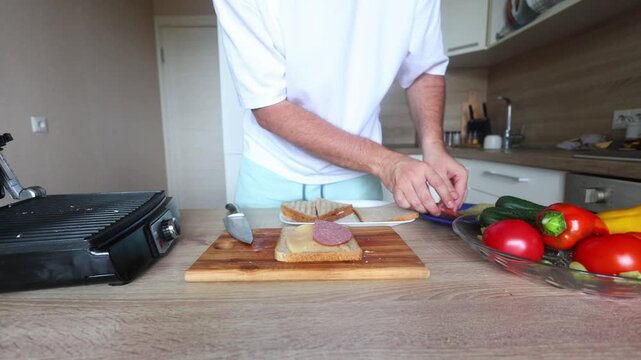 Man assembles and cuts a cheese salami sandwich on whole grain bread, grill, fresh veggies, and ingredients visible in a bright, modern kitchen.