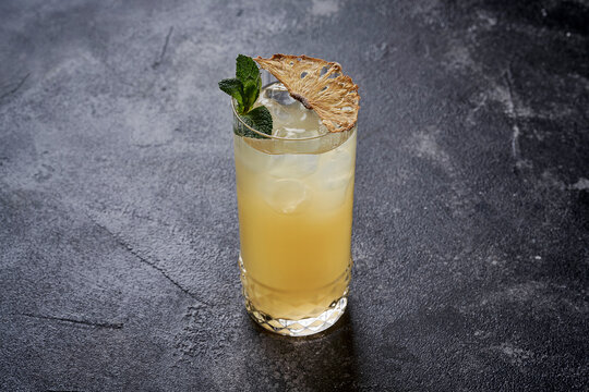 Pineapple cocktail with mint garnish and dried pineapple slice in a highball glass on a dark stone background.