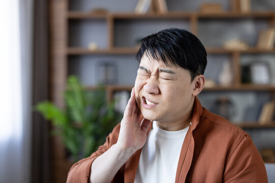 Asian man experiencing intense oral discomfort, clenching his jaw and holding his cheek to alleviate the throbbing sensation from a sensitive tooth, indicating a need for urgent dental care