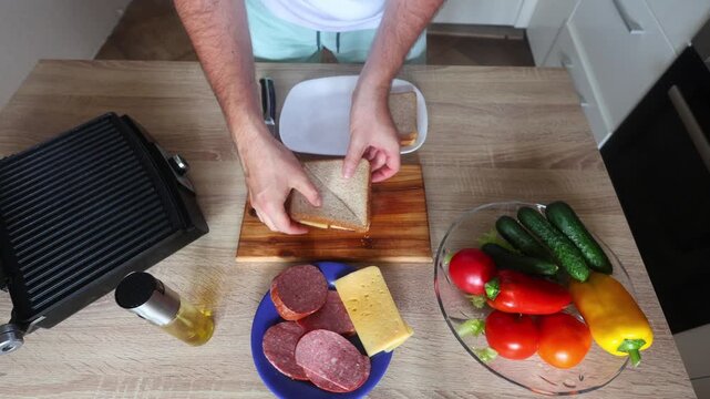 Man oils and grills a cheese salami sandwich on a panini press, top down view with fresh veggies, bread, and ingredients neatly arranged on a wooden counter.