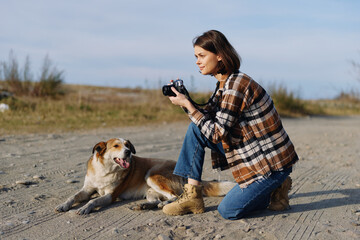 Obraz premium woman photographer kneels on sandy outdoor terrain with a dog beside her holding a camera and focusing on subjects in the open landscape under a clear sky
