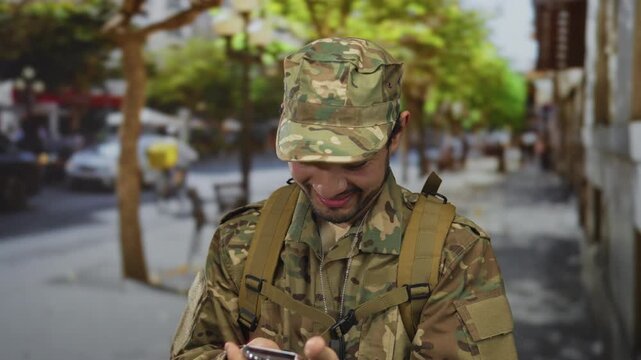 Man soldier in camouflage uniform sticking out tongue and flashing v sign while taking a selfie on a city street; joy.