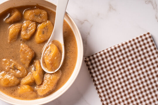 Top view of Kolak pisang on a white plate with a white spoon. Indonesian dish for breaking the fast, during Ramadan or Eid al-Fitr. Sweet soup made from bananas with coconut milk and brown sugar. 