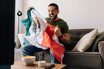 Happy Mexican soccer supporter celebrating at home while holding the national flag. Adult man enjoying the match on TV with snacks and drinks in a cozy living room. © kleberpicui