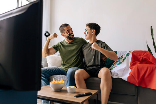 Mexican father and teenage son celebrating a football moment together at home. Happy family supporters cheering on the couch with national flag, snacks and drinks.
