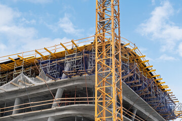 Closeup of upper floors of curved concrete building under construction with yellow tower crane, dense scaffolding and protective netting against sky   © Nannie