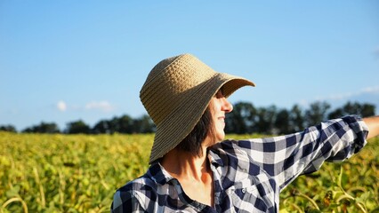 Happy smiling female farmer looks into sunflower field and enjoys harvest. Portrait of adult beautiful agronomist in straw hat with yellow plantation at background. Concept of agricultural business