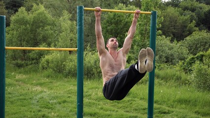 Athletic man trains abdominal muscles raising legs on horizontal bar at sports ground. Young sportsman exercising on pull-up bar outdoor. Sporty athlete working out at nature. Healthy lifestyle