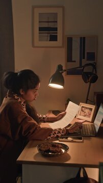Vertical shot of young adult multiethnic woman typing on laptop keyboard entering data while analyzing document, remote employee working at cozy desk with lamp in home office