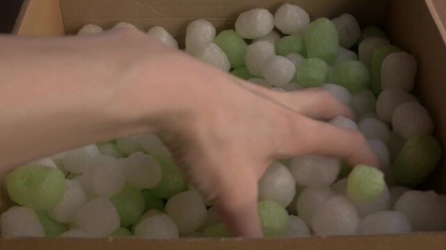 Hand reaches into a box filled with biodegradable starch foam peanuts in green and white, showcasing eco-friendly packaging materials. Ecofriendly packaging