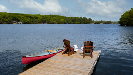 Obraz premium Elevated wide-angle shot of a wooden dock on a calm lake with two Adirondack chairs and striped towels. A red canoe tied to the pier adds adventure to this tranquil Muskoka summer nature getaway.