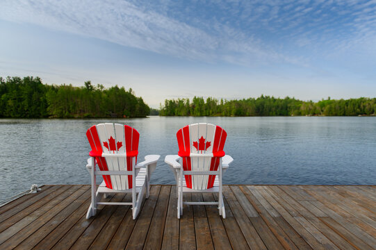 Wide-angle view of two white Adirondack chairs with red maple leaf flags on a rustic dock. Calm water and forest evoke a serene Canadian lake house experience focused on patriotism and relaxation.
