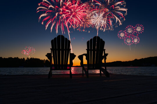 Night scene in Muskoka with two Adirondack chairs silhouetted on a dock. Fireworks burst over a calm lake, celebrating Canada Day at a summer cottage. Ideal for festive and seasonal holiday themes.