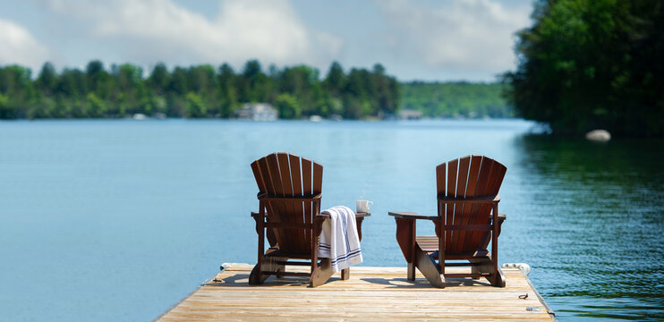 Wide-angle view of a wooden dock with two Adirondack chairs facing a calm lake. A striped towel suggests a relaxing morning. Lush forest and bright sky create a peaceful atmosphere with copy space.