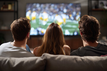 Naklejka premium Back view of three excited friends watching the Super Bowl championship on TV while sitting on a couch at home 