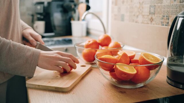 Woman in light sweater slices oranges on wooden cutting board in kitchen, with bowls of oranges and kitchen appliances visible in background, showcasing continuous preparation process