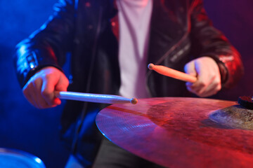 Obraz premium Man with drumsticks playing cymbal in color lights against dark background, closeup