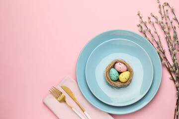 Fototapeta premium Easter celebration. Festive table setting with painted eggs, willow branches and cutlery on pink background, flat lay with space for text