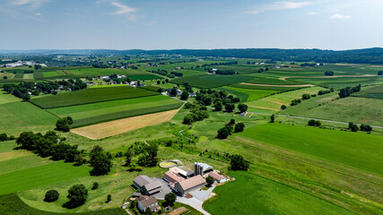 Aerial view of a rural area showing fields, farms, and a clear blue sky. Green fields stretch across the landscape. Buildings are visible near a dirt road. © Greg Kelton