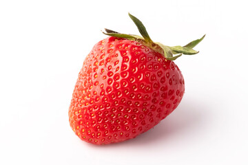 A ripe strawberry sits on a white surface. The fruit shows bright red color with tiny seeds visible all over. Green leaves are on top, indicating freshness