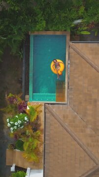 Aerial view of a woman enjoying her vacation floating on a yellow inflatable ring in a swimming pool at a tropical luxury villa