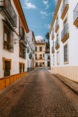 Obraz premium Cobblestone street winding through the historic old town of Cordoba, Spain, with traditional white buildings displaying yellow details and ornate balconies under a clear blue sky