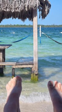Beach vacation POV showing sandy toes in foreground with crystal clear tropical lagoon, buoys and wooden platforms extending into emerald waters.