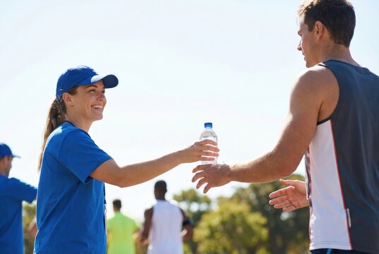 A smiling female volunteer in a blue shirt and cap hands a bottle of water to a male runner during a marathon or outdoor sporting event.