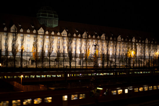 Two night trains (both lit) passing in front of a long row of trees and identical buildings, also well lit
