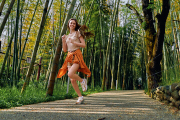 Obraz premium Woman running in bamboo forest path, smiling and wearing orange skirt and sneakers, outdoor jog for fitness and travel, healthy young adult enjoying nature and sunlight on gravel trail