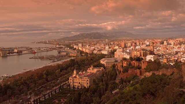 A warm, orange sunrise over a bustling coastal Malaga city, calm harbor with ships, palm-lined promenade, and historic center beneath a skyline of modern buildings and distant mountains. Malaga, Spain