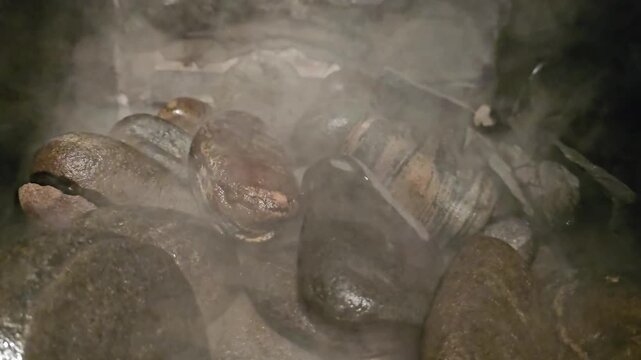 Close-up view of round, heated stones in a sauna heater. Water evaporates from the wet surfaces, generating dense steam that rises in the dim, humid environment. Captures the heat and moisture typical